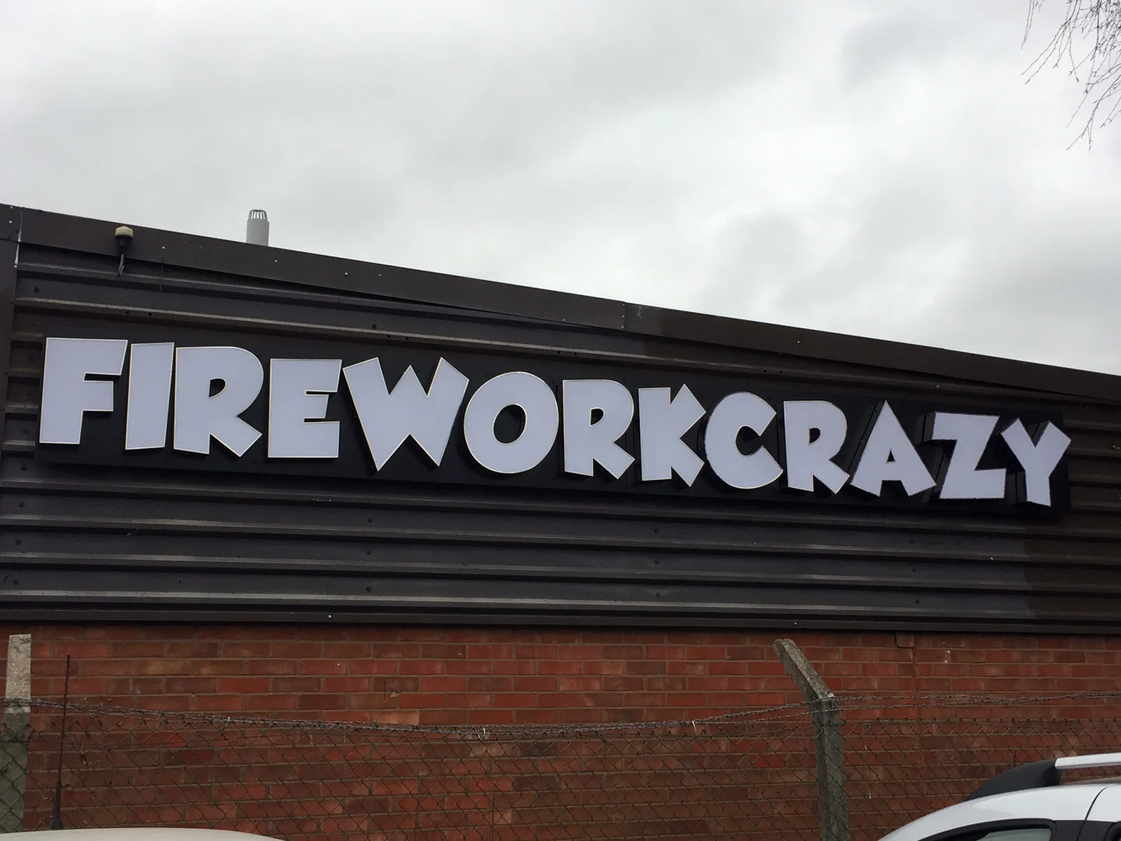 White FIREWORKCRAZY letters mounted on a dark exterior sign band above brickwork