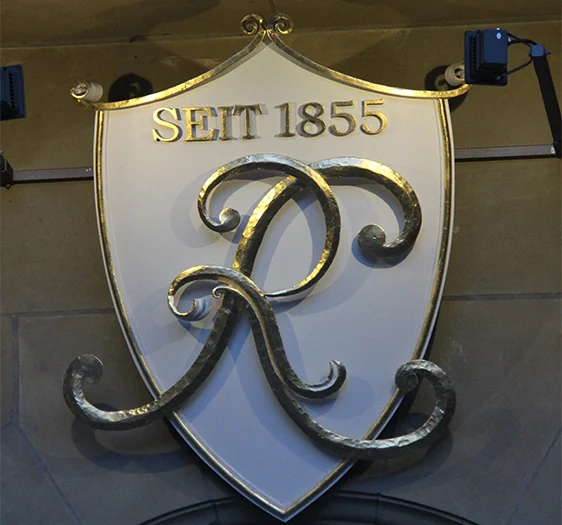 A shield-shaped plaque with raised decorative lettering and a reflective ornamental monogram.