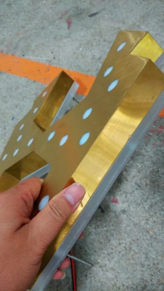 A hand holding a gold-colored sign component with cut-outs, deep sides and a projecting fixing rod.