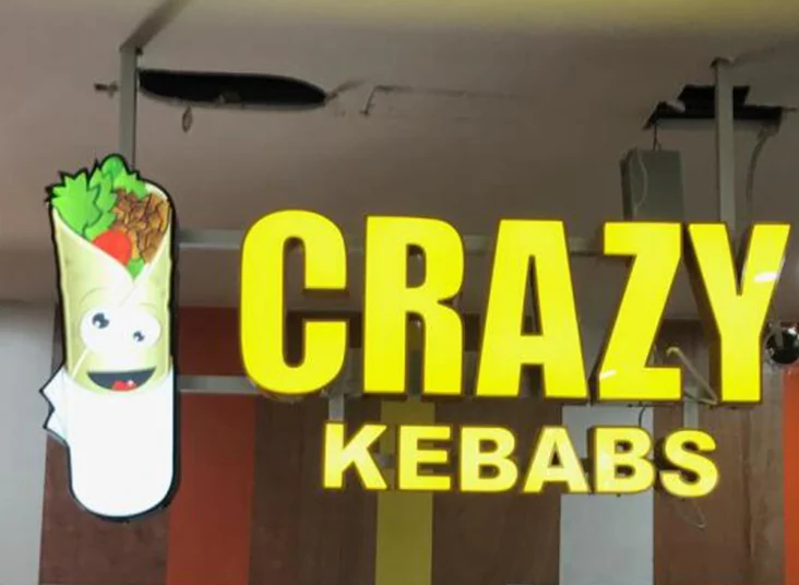 A colorful food-shaped logo beside yellow illuminated lettering at a commercial counter.