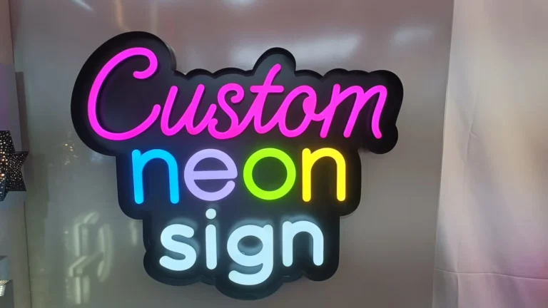 A three-line custom neon sign sample with pink, blue and yellow lettering on a dark shaped backing.