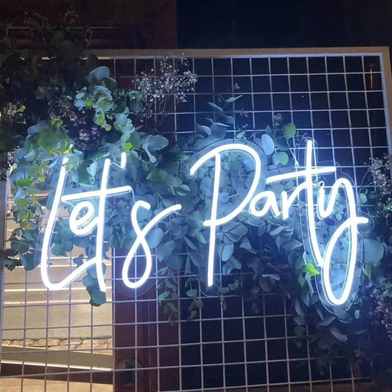 White illuminated script lettering on clear shaped backing in front of a metal grid and foliage.