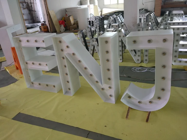 Unlit white ENJ letter bodies with raised returns and rows of clear bulbs in a workshop.
