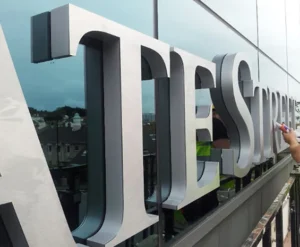 Large silver-colored dimensional letters viewed from the side beside a glass building facade.