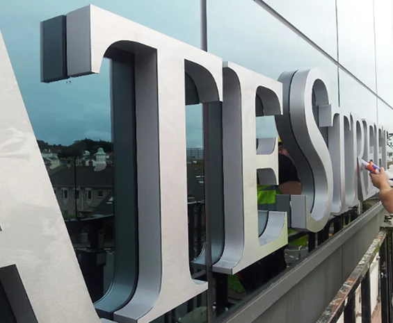 Large silver-colored dimensional letters viewed from the side beside a glass building facade.