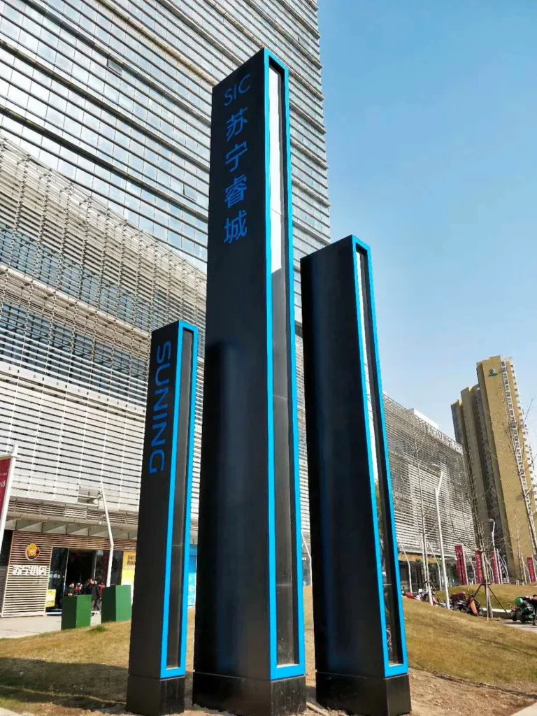 Tall dark freestanding sign structures with blue illuminated lettering beside a modern building.