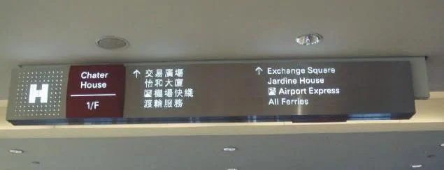 An overhead directional panel with bilingual destinations, arrows and transport symbols.