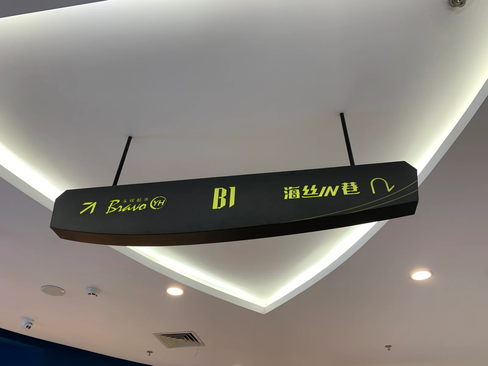 A dark directional sign suspended from an interior ceiling, with destination text and arrows.